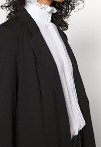 Musta bleiseri terävillä läpäillä valkoisen, tekstuurilla varustetun paidan päällä, jossa on röyhelökaulus ja napitus. Lähikuva kerroksittaisista vaatteista.