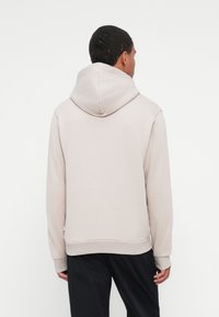 Felpa beige chiaro con cappuccio regolabile, polsini e orlo a costine, texture morbida e vestibilità rilassata. Indossata su pantaloni scuri.