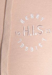 Tessuto di cotone rosa chiaro con logo circolare ricamato contenente "H.I.S" e numeri, affiancato da un accento texturizzato e intrecciato sul bordo.
