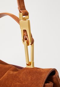 Brown suede handbag with a gold-tone metal clip, featuring a textured finish and a structured shape. Accent details on the strap.