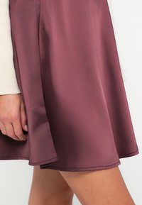 Burgundi szatén szoknya lágy szabással, sima textúrával és enyhe lefele bővüléssel az alján, világos színű toppal párosítva.