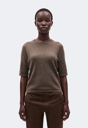 Jeune femme à la peau foncée aux cheveux courts portant un pull en maille marron à manches courtes et un pantalon marron texturé assorti, debout immobile.