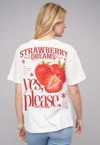 T-shirt en coton blanc avec un graphique de fraise rouge audacieux et le texte "RÊVES DE FRAISE oui, s'il vous plaît." Manches courtes, coupe décontractée.