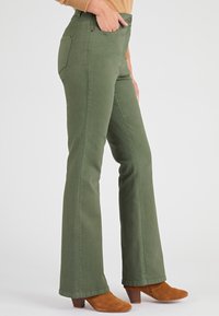 Olijfgroene flared jeans met een gladde textuur, voorzien van een knoopsluiting, voor- en achterzakken, en gecombineerd met bruine hoge laarzen.