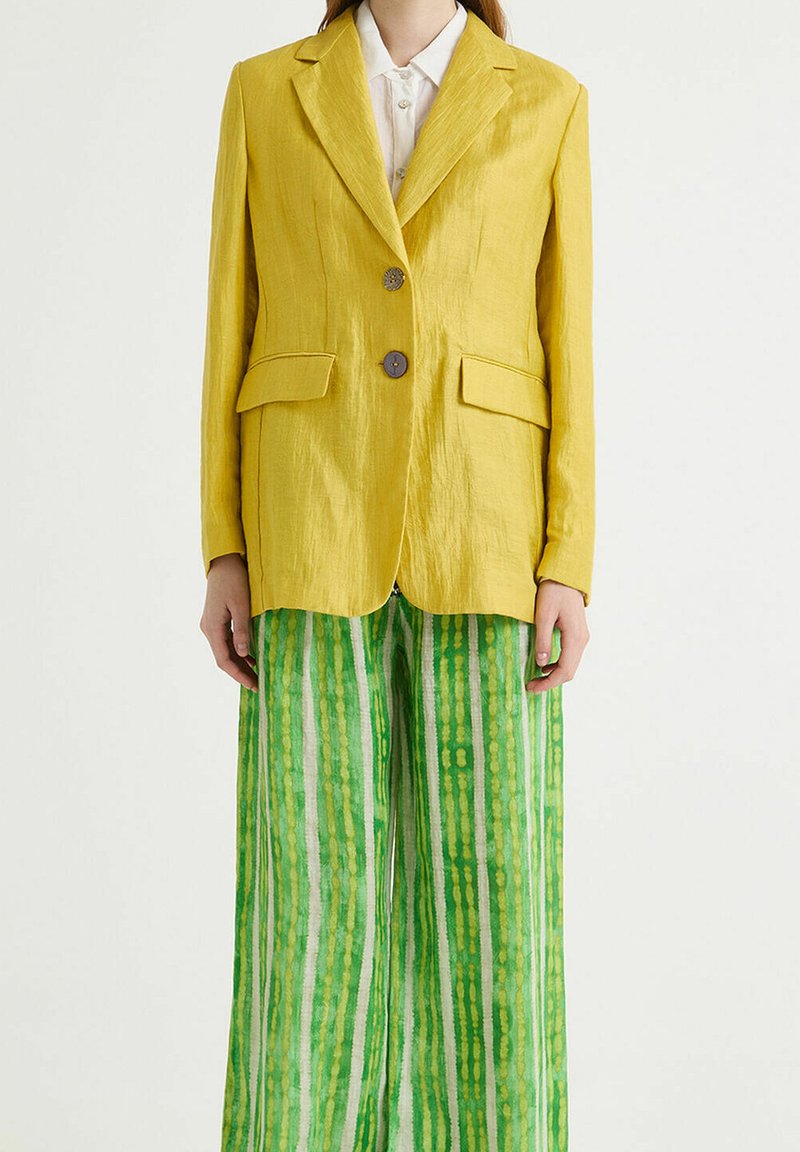 Blazer en lin jaune avec deux boutons, revers crantés et poches. Associé à un pantalon large à rayures verticales vertes.