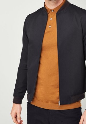 Homme portant une veste noire zippée sur un polo marron à boutons, avec un pantalon noir, debout devant un fond clair uni.