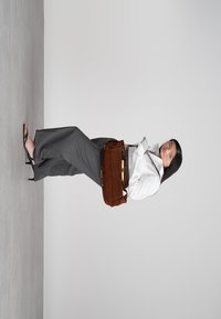 Nainen, jolla on päällään valkoinen paita ja harmaat housut, kantaa ruskeaa mokkaista olkalaukkua, seisoo harmaalla lattialla yksinkertaista valkoista taustaa vasten.