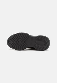 Semelle de chaussure noire avec des motifs de bande de roulement texturés comportant des rainures courbes et droites pour l'adhérence, présentée sur un fond blanc uni.