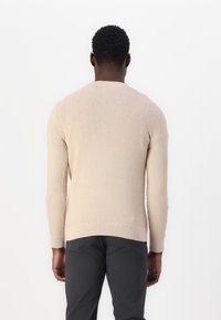 Beige gebreide trui met een gestructureerd ruitpatroon, geribde manchetten en zoom, gecombineerd met donkergrijze broek. Achteraanzicht weergegeven.