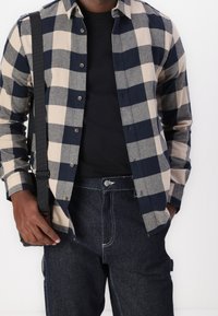 Man in een zwarte shirt en een beige-blauw geblokte overhemd, donkere spijkerbroek en draagt een zwarte schoudertas.