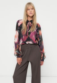 Flerfarget bluse med abstrakt rosa, lilla og svart mønster. Har volangermodellerte ermer og en knyttet sløyfe i halsen, sammen med grå stripete bukser.