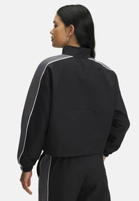 Veste noire raccourcie avec accents gris et passepoil blanc. Dotée d'un col montant et de poignets élastiques. Tissu lisse avec une coupe décontractée.