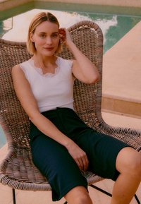 Femme aux cheveux blonds courts portant un haut blanc sans manches et un short noir, assise sur une chaise en osier près d'une piscine.