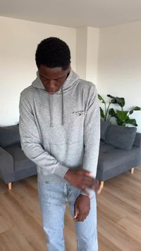 Νέος άνδρας με γκρι φούτερ ρυθμίζει το μανίκι του, στέκεται στο σαλόνι με γκρι καναπέ και πράσινα φυτά στο φόντο.