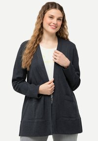 Mörkgrå, öppen cardigan med lång ärm, ribbad textur, två framfickor och avslappnad passform, bärs över en ljus skjorta.