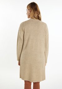 Beige pletený kardigan s dlouhými rukávy, rovného střihu a bočními rozparky. Měkká textura a jednoduchý design bez dalších ozdob.