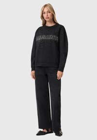 Czarna bluza z napisem "ALLSAINTS" ozdobionym cekinami, dekolt okrągły, luźny krój, w połączeniu z luźnymi czarnymi spodniami i płaskimi butami.