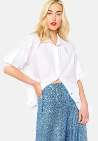 Camicia bianca oversize a maniche corte, con apertura a bottoni sul davanti. Abbinata a una gonna blu scintillante in paillettes che brilla.