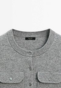 Cardigan gris en tricot avec encolure ronde, quatre boutons gris sur le devant, et deux poches poitrine à boutons.