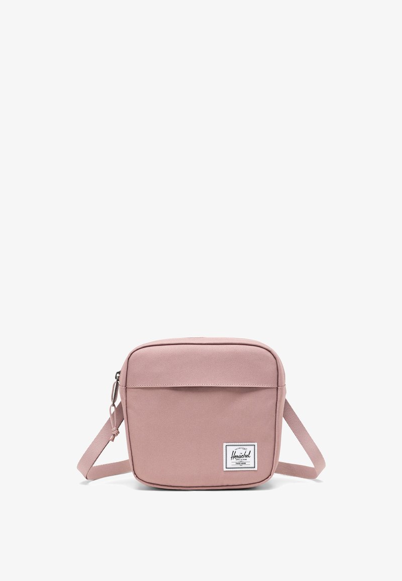 Čtvercová růžová crossbody taška vyrobená z odolné látky, obsahující přední kapsu, nastavitelné rameno a nášivku s logem v pravém dolním rohu.