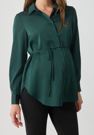 Vrouw draagt een lange mouw donkergroene zwangerschapsblouse met knopen en een strikceintuur, gecombineerd met een zwarte broek, tegen een effen achtergrond.