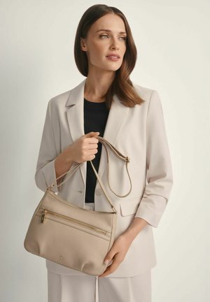 Kobieta w beżowym garniturze trzyma beżową skórzaną torebkę z złotym suwakiem, spoglądając na bok na tle jednolitego jasnego tła.