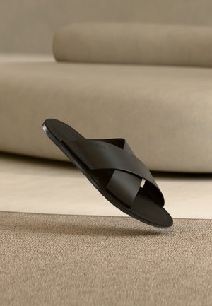 Čierna nazúvacia sandála s prekríženými remienkami vznášajúca sa nad béžovým kobercom pred zaoblenou béžovou pohovkou.