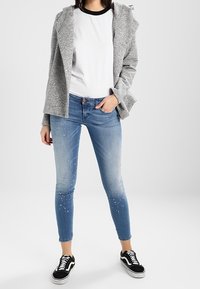 Grå hoodie med knappar, vit t-shirt, slitna blå skinny jeans och svarta sneakers med vita sulor. Sammanfattningsvis en avslappnad outfit.