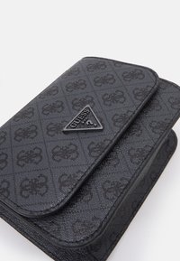 Czarny portfel z tworzywa sztucznego o teksturowanej powierzchni, z powtarzającym się wzorem i trójkątnym logo Guess z przodu. Projekt z klapką.