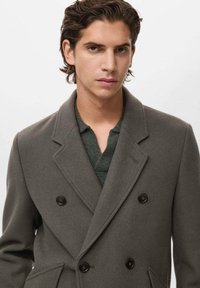 Cappotto grigio in lana doppiopetto con bottoni neri, revers a rovescio e tasche laterali, indossato sopra una camicia texture verde scuro.