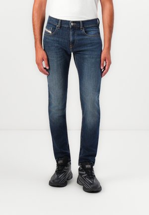 Homme portant un jean slim bleu et des baskets Diesel noires, debout devant un fond clair uni.