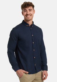 Lächelnder junger Mann mit lockigem Haar, der ein marineblaues Hemd mit Knöpfen und beige Hosen trägt und mit einer Hand in der Tasche steht.