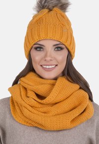 Neulepipo ja infinity-huivi kirkkaassa sinapinkeltaisessa värissä. Pipossa on teksturoitu kuvio ja pörröinen pom-pom. Molemmat ovat pehmeitä ja mukavia.