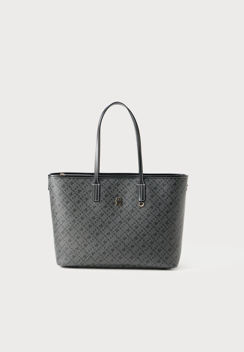 Svart totebag med monogrammønster, doble skulderstropper, detaljer i gulltone med logo og ring, stående oppreist mot en hvit bakgrunn.