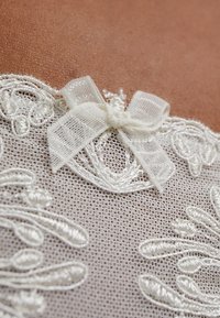 Dentelle blanche avec une broderie florale intricate et un petit nœud en tulle délicat. La texture semble douce et légère.