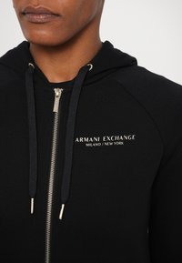 Crni dukserica sa zatvaračem i zlatnim detaljima, s izvezenim logotipom "Armani Exchange Milano/New York" na prsima i teksturiranim materijalom.