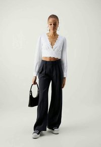 Blusa branca curta com padrão de ilhós, mangas longas e decote em V; calças escuras de cintura alta; bolsa preta; ténis preto e branco.
