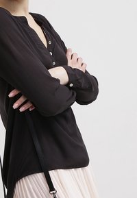 Blusa nera con bottoni a camicia e maniche lunghe, con scollo rotondo e bottoni scuri, abbinata a una leggera gonna plissettata e una cintura nera.