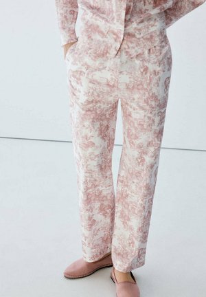 Persona con pantalones estampados rosa claro y chaqueta a juego, de pie con las manos en los bolsillos y calzando zapatos rosa claro sin cordones.