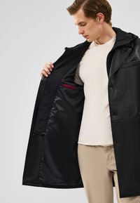 Zwarte leren jas met een capuchon en knoopsluiting, voorzien van een interieur gevoerd met glad zwart stof en een klein rood pocketdetail.
