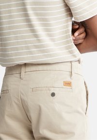 Beiga byxor i lätt tyg, med en bakficka med knapp och en Timberland-logotyp på midjan.