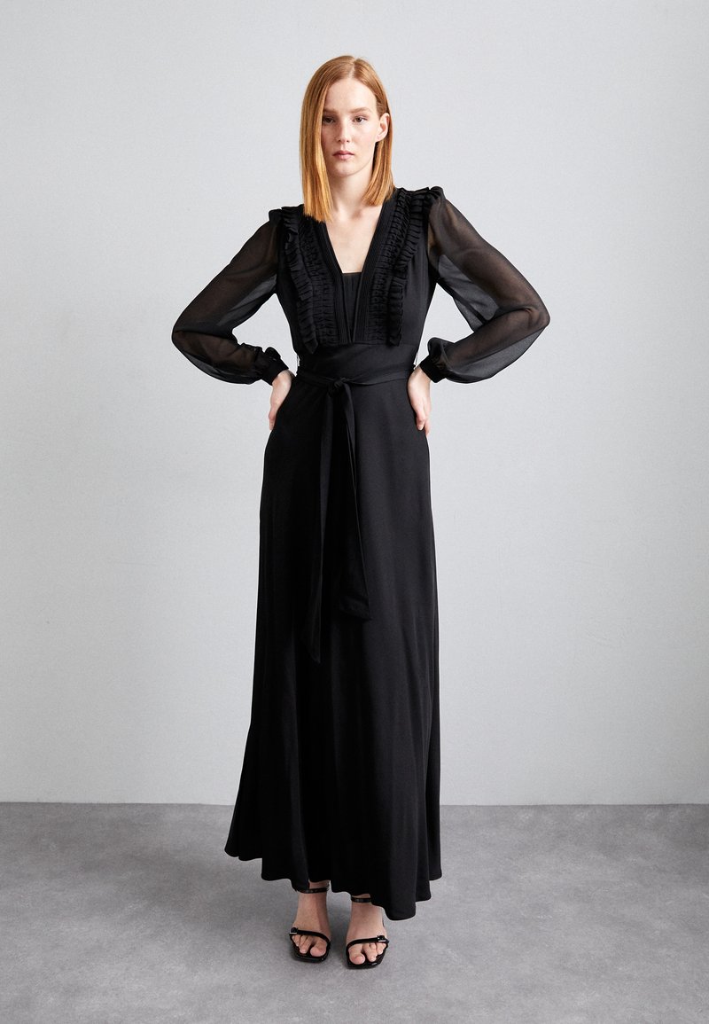 Robe maxi noire avec un décolleté en V, des accents en volants et de longues manches transparentes. Nouée à la taille, le tissu fluide tombe élégamment jusqu'au sol.