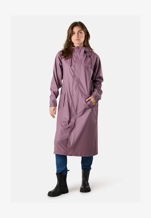 Frau steht und trägt einen langen mauvefarbenen Regenmantel mit Kapuze, blaue Jeans und schwarze Gummistiefel, mit einer Hand in der Tasche.