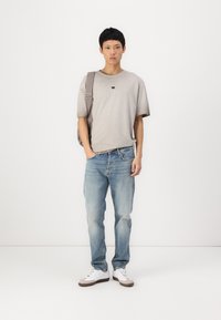 T-shirt gris à manches courtes avec logo, jean en denim bleu clair, baskets blanches avec accents gris, et sac à bandoulière gris. Tenue simple et décontractée.