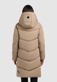 Beige puffertakaró kapucnival, chevron mintával, hátul gombos záródással, és kis márkalogóval az alján. Simá, steppelt textúra.