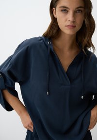Pullover blu navy con cappuccio, caratterizzato da dettagli in metallo argentato, tessuto testurizzato e una vestibilità relaxed con maniche arrotolate.