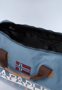 Borsa in tessuto blu con manici marroni, riportante un patch della bandiera islandese e un interno nero con cerniera. Superficie esterna testurizzata.
