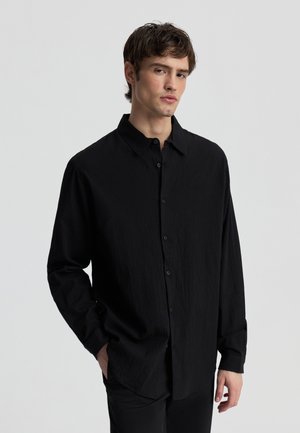 Giovane uomo con capelli scuri corti, che indossa una camicia nera a maniche lunghe e sloose con bottoni e pantaloni neri, in piedi davanti a uno sfondo chiaro e uniforme.