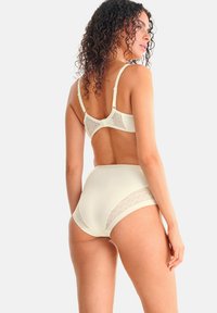 Femme aux cheveux bouclés portant un soutien-gorge couleur crème et des culottes taille haute avec des détails en maille, vue de dos sur un fond uni.