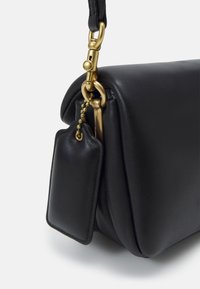 Czarna skórzana torebka o gładkiej fakturze, z zapięciem z półprzezroczystego złotego metalu i zwisającą metką na łańcuszku. O zaokrąglonym kształcie.